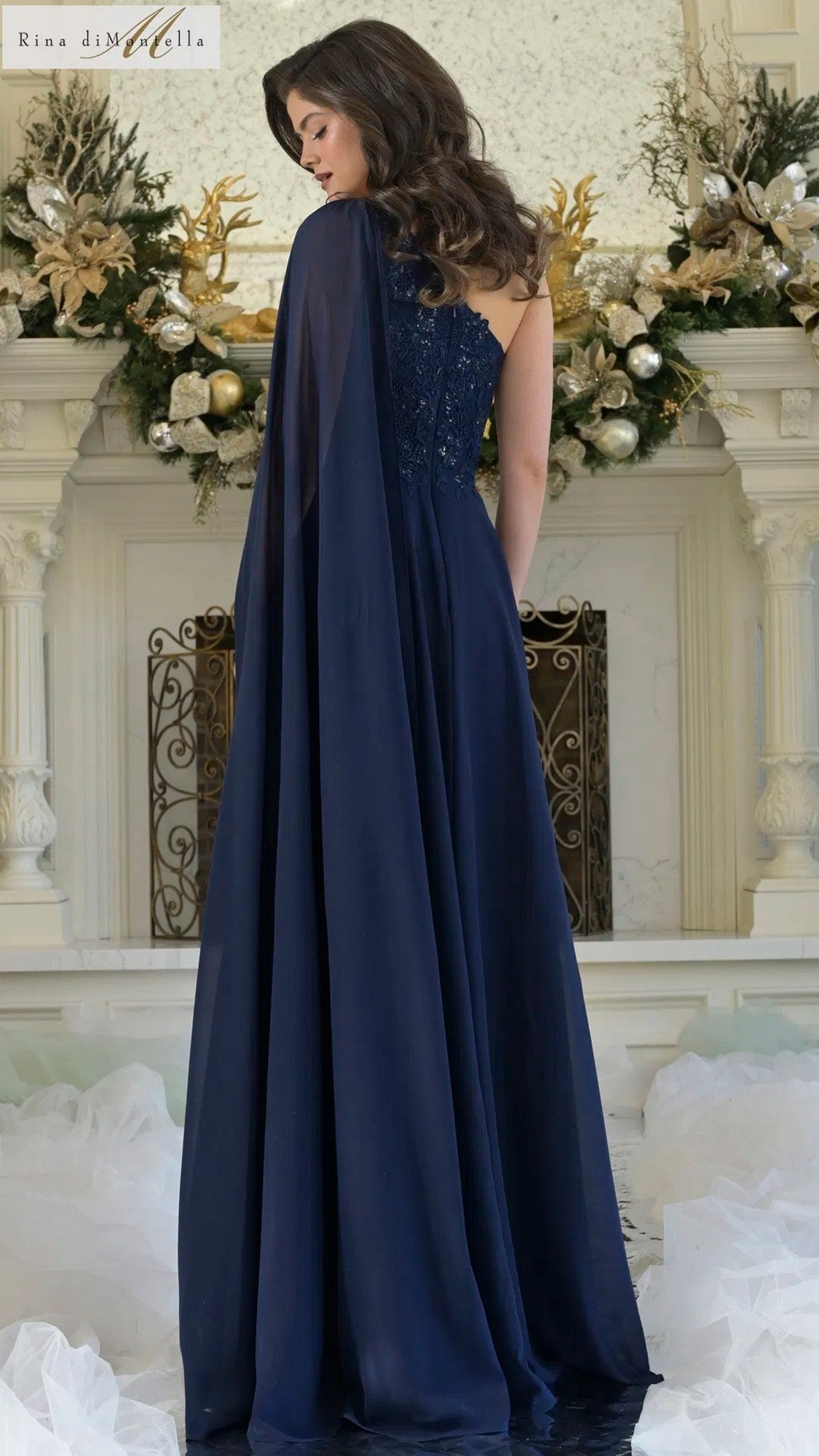 A woman faces away from the camera, wearing the Rina Di Montella RD3354 long formal dress with a dark blue, floral-embroidered bodice and flowing cape, standing before a festively decorated white fireplace.